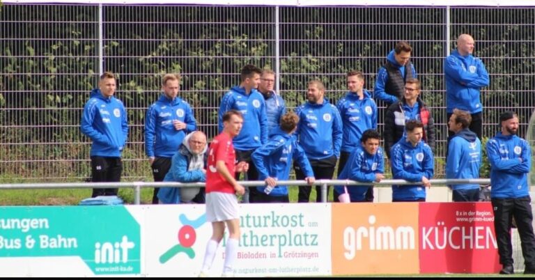 Szene am Spielfeldrand: mehrere Personen in blauer Vereinskleidung beobachten ein Fußballspiel